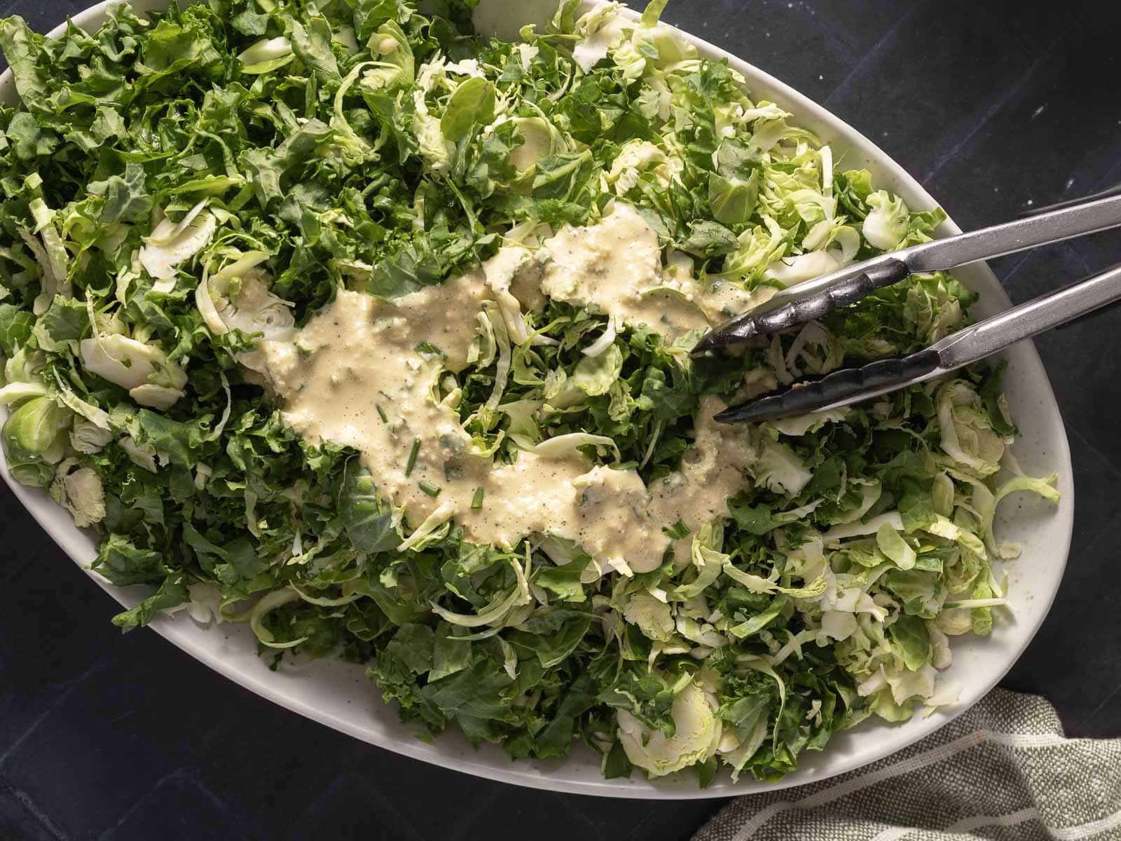 A bowl of chopped green kale salad with dressing, accompanied by metal tongs on a dark surface.