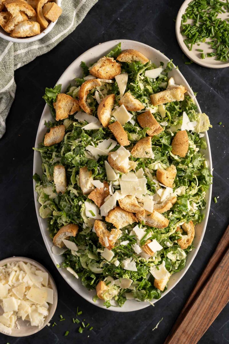 A large oval plate of kale and Brussels sprout salad topped with croutons and shaved Parmesan cheese, surrounded by small bowls of extra Parmesan and chopped herbs.