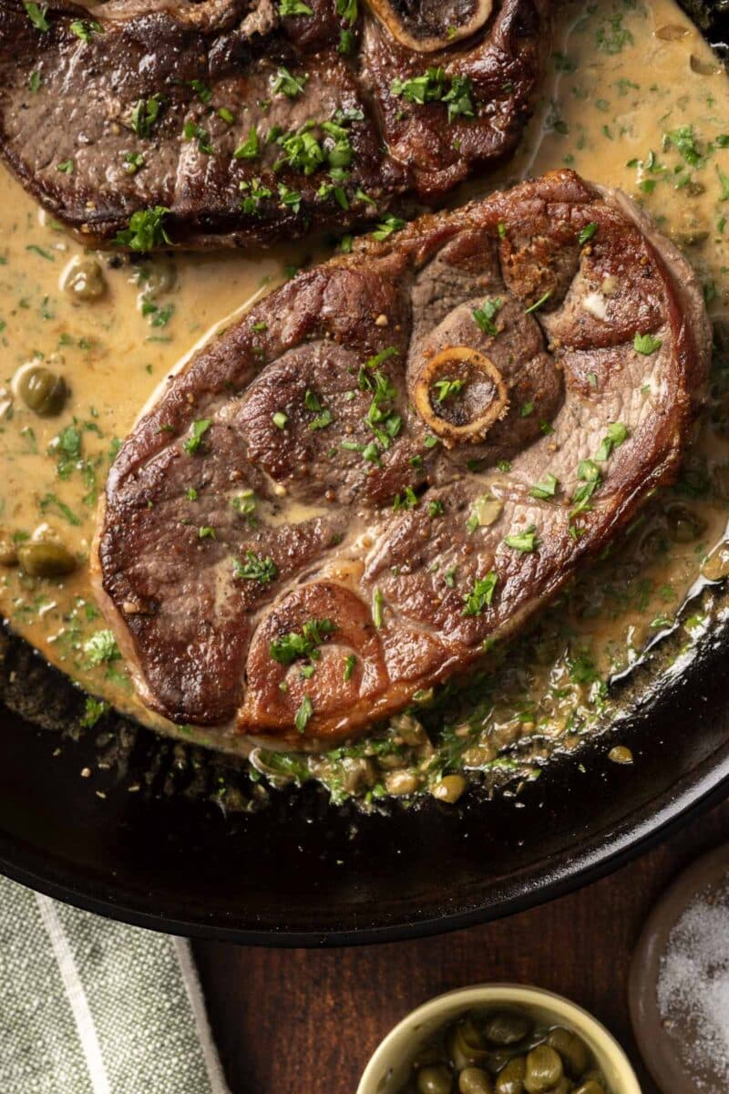 Two cooked osso buco pieces in a creamy herb sauce, served in a pan like a delightful lamb steak recipe. A small bowl of capers sits beside the pan.