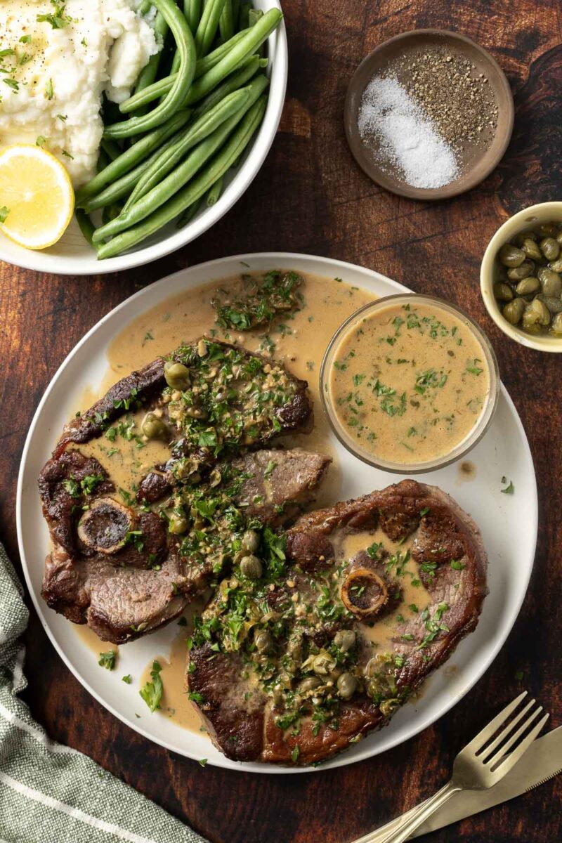 A plate with two pieces of beef in caper sauce, accompanied by a small dish of extra sauce. Green beans, mashed potatoes, and a lemon wedge are visible in the background, along with salt, pepper, and capers—a perfect inspiration for your next lamb steak recipe.