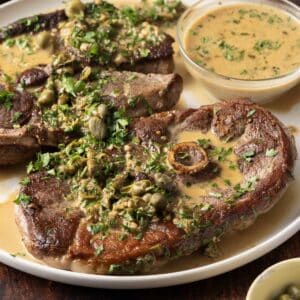 Plate of cooked lamb chops garnished with capers and green herbs, served with a creamy mustard sauce on the side—an exquisite lamb steak recipe perfect for any special occasion.