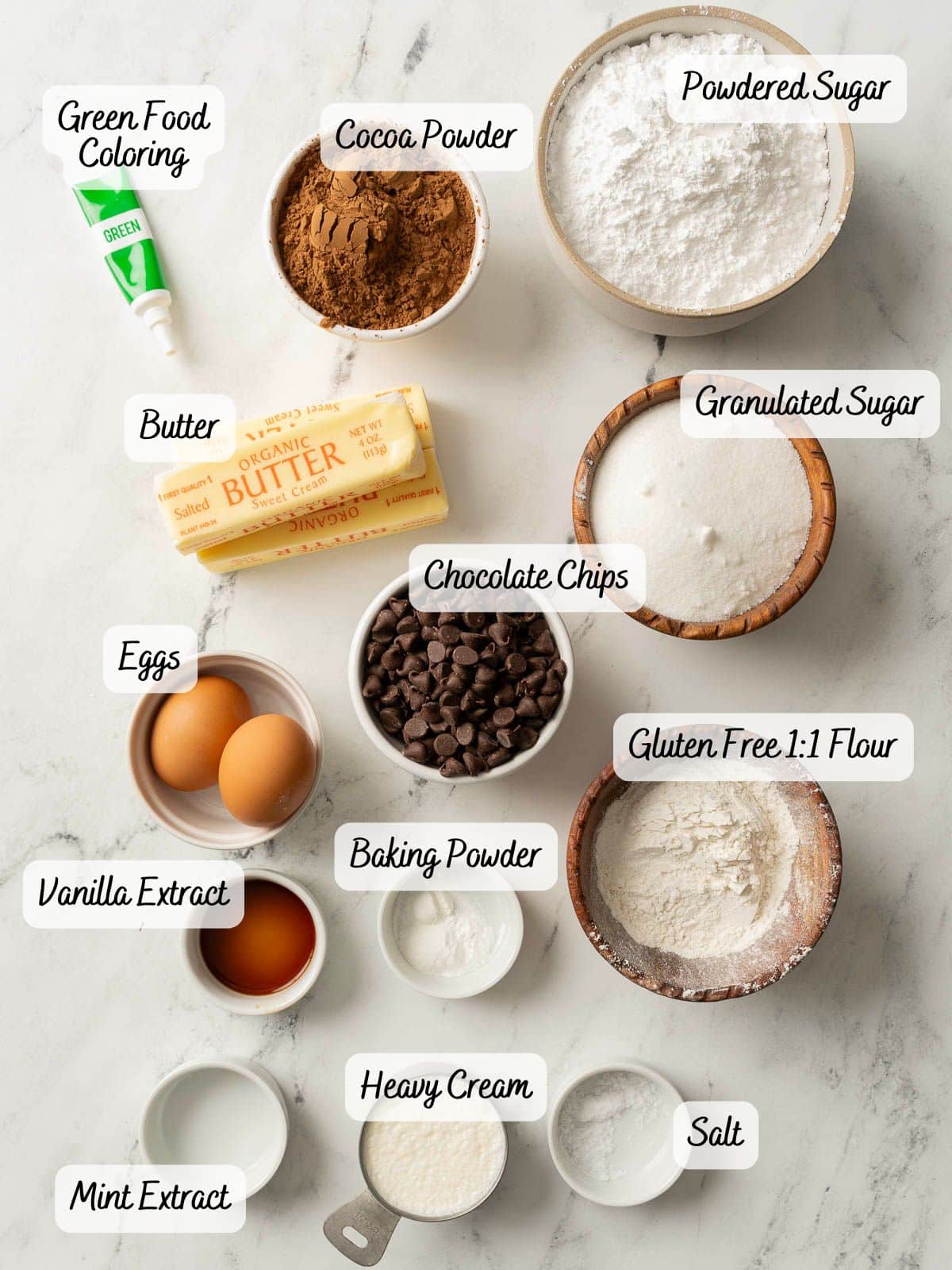 Top view of baking ingredients on a marble surface, labeled and ready for gluten-free mint chocolate brownies—featuring flour, sugar, cocoa powder, butter, eggs, chocolate chips, and more.