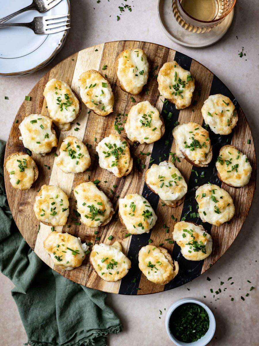 A round wooden board with several cheesy bread bites garnished with chopped herbs and a hint of Parmesan. Plates, forks, and a glass are visible nearby.