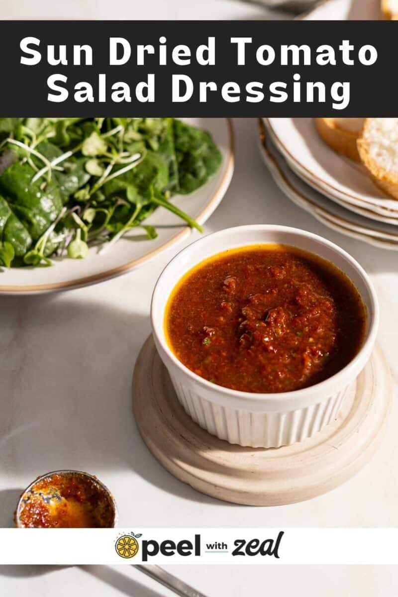 A bowl of sun-dried tomato salad dressing on a table beside a salad and a slice of bread.