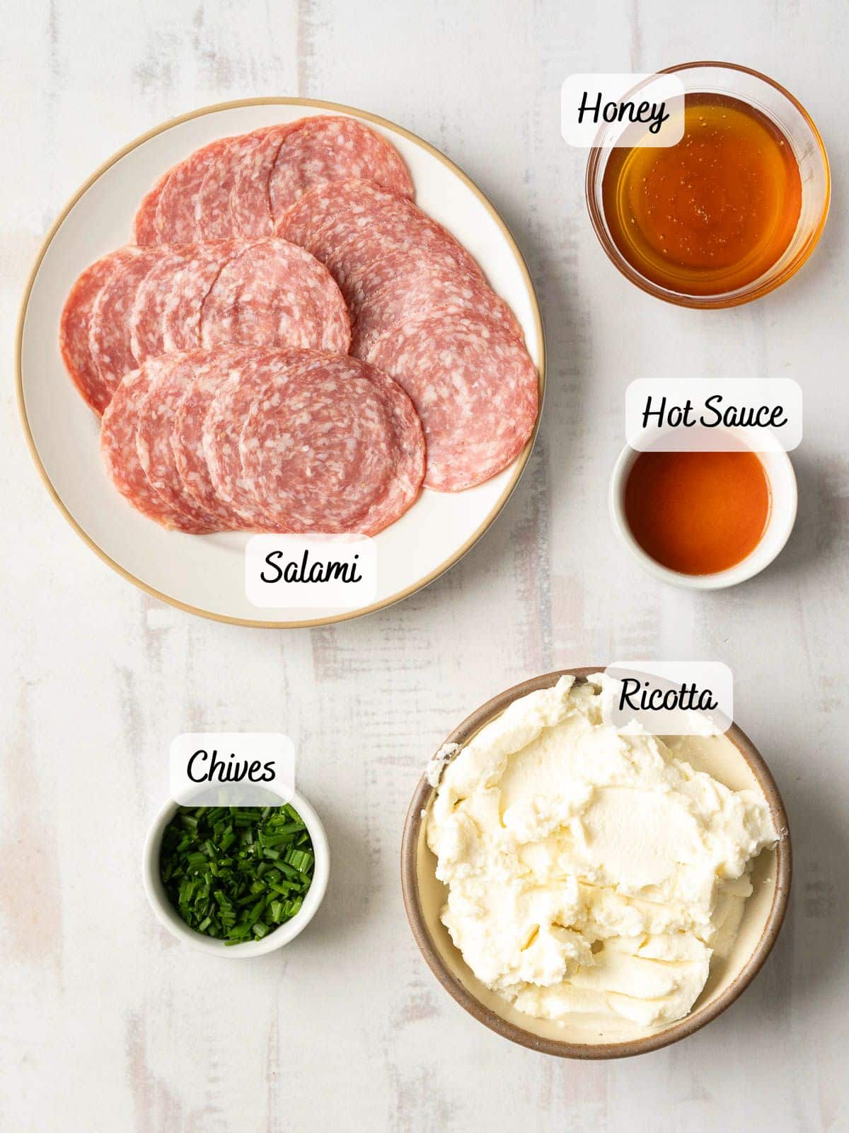 A plate of sliced salami, bowls of honey, hot sauce, ricotta, and chopped chives are arranged on a light surface.