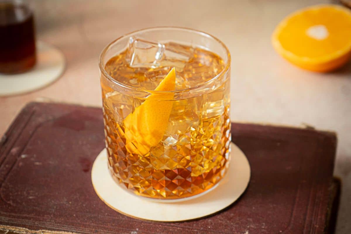 A glass of amber-colored autumn cocktail with ice and an orange peel garnish sits on a coaster, with a halved orange and another glass blurred in the background-perfect for those who love fall cocktails.