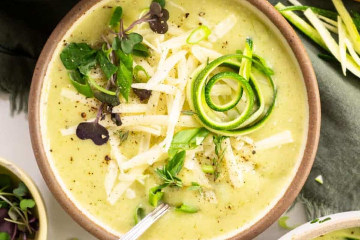 A bowl of creamy soup garnished with shredded cheese, zucchini spirals, and fresh greens. A metal spoon is partially visible in the soup.