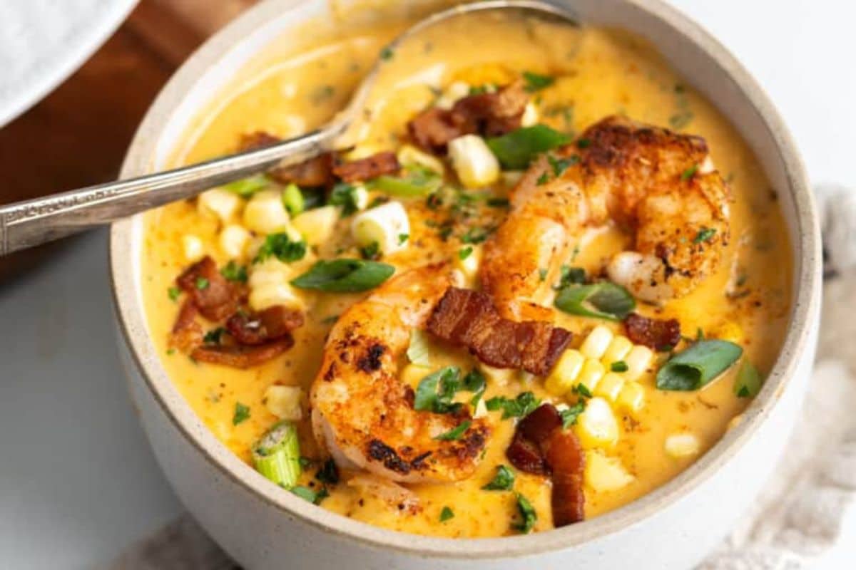A bowl of shrimp chowder with grilled shrimp, corn, bacon, and green onions, garnished with herbs. A spoon rests in the bowl.