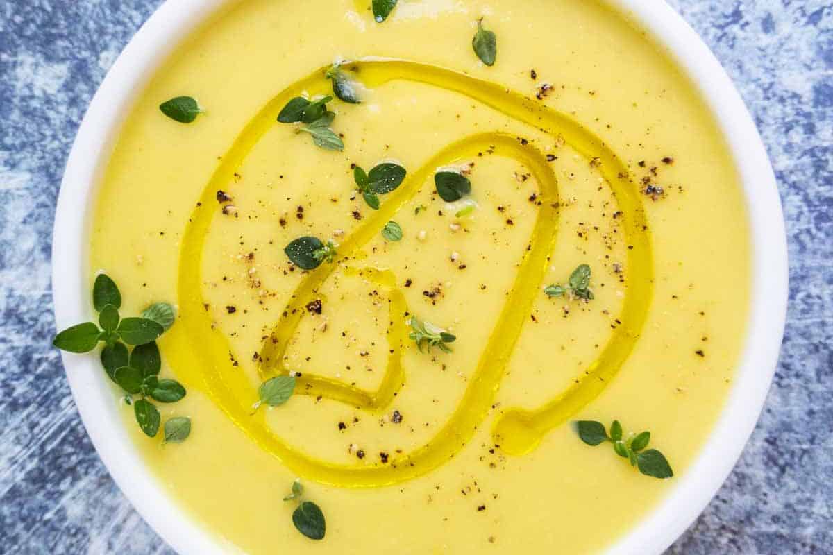 A bowl of creamy yellow soup garnished with a swirl of olive oil, black pepper, and fresh herbs.