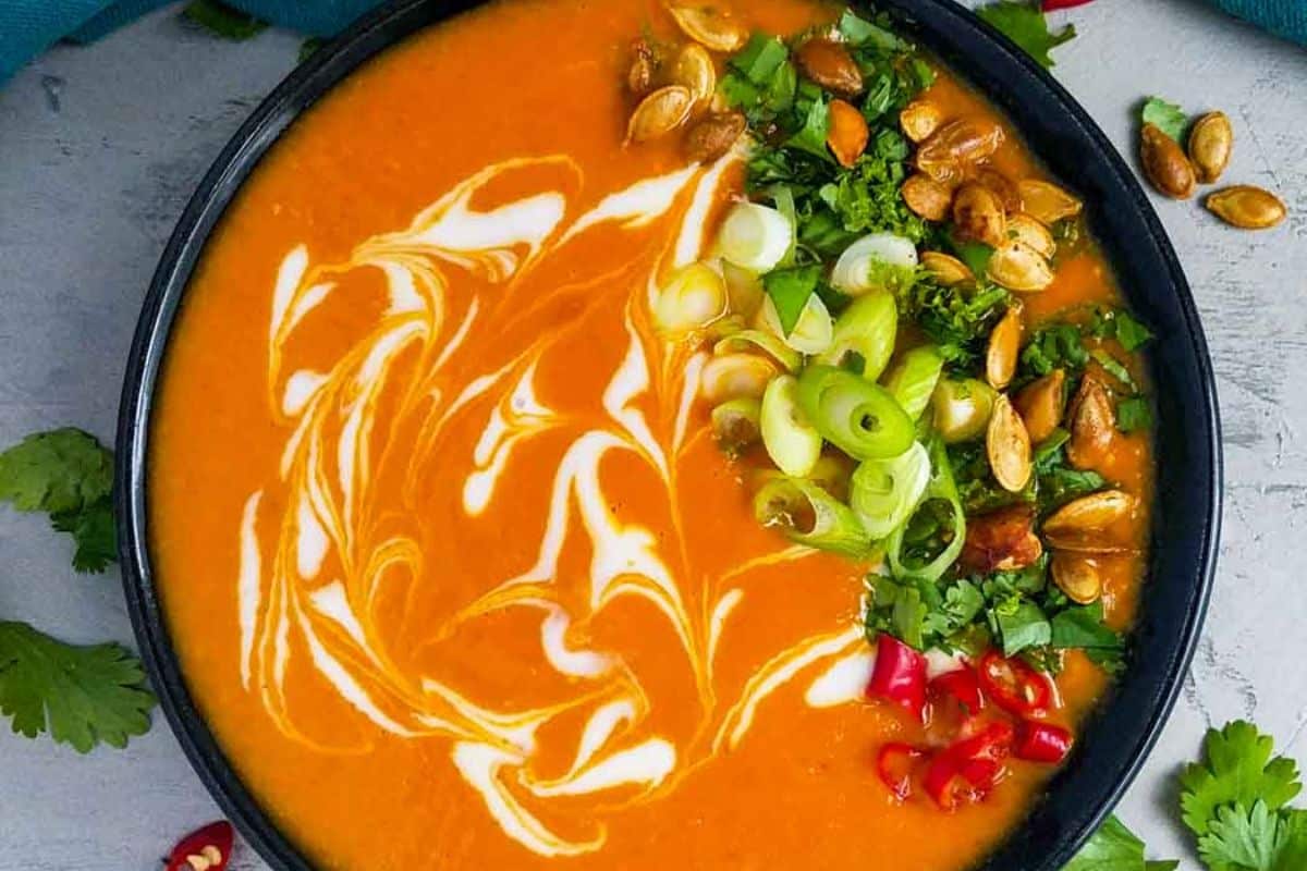 A bowl of creamy orange soup garnished with fresh herbs, sliced green onions, roasted seeds, and a swirl of cream.
