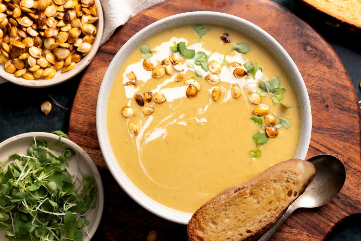 A bowl of creamy soup garnished with seeds, microgreens, and a drizzle of cream. There's a slice of toasted bread on the side, with another bowl of seeds and microgreens nearby.