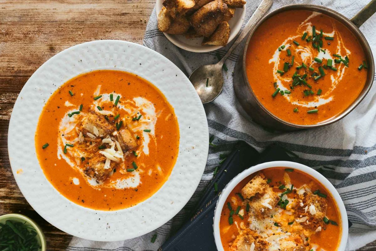 Three bowls of creamy tomato soup garnished with cream, herbs, and croutons are displayed on a wooden table. A spoon is placed beside the pot.