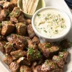 A platter of steak bites garnished with herbs sits next to a bowl of rich blue cheese sauce, making an irresistible appetizer, with toothpicks neatly arranged in a cup.