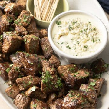 A platter of steak bites garnished with herbs sits next to a bowl of rich blue cheese sauce, making an irresistible appetizer, with toothpicks neatly arranged in a cup.
