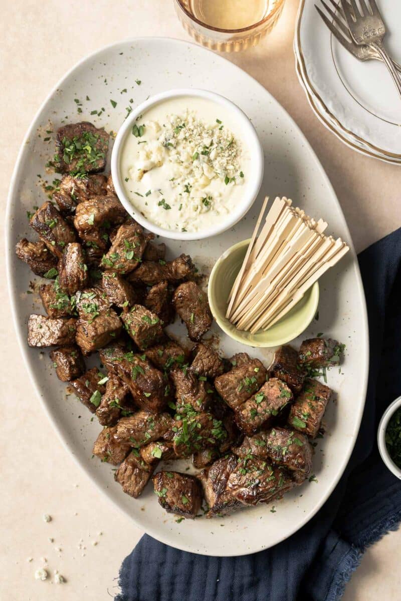 A plate of succulent steak bites garnished with herbs is served alongside a bowl of dipping sauce and toothpicks, elegantly arranged on a table with wine glasses and a napkin, making for the perfect appetizer.