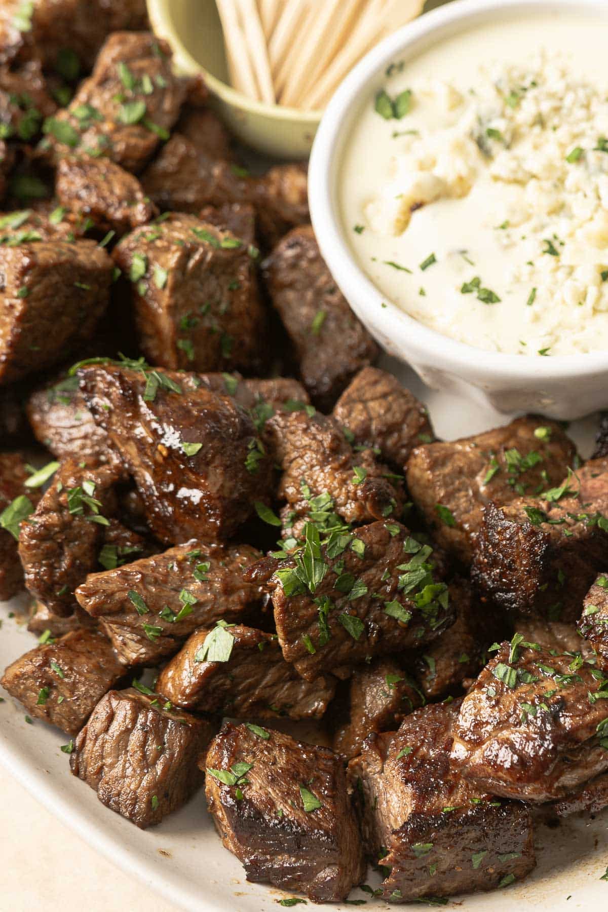 Close-up of succulent steak bites, an irresistible appetizer, garnished with parsley and served on a plate with a side of creamy blue cheese sauce and toothpicks.