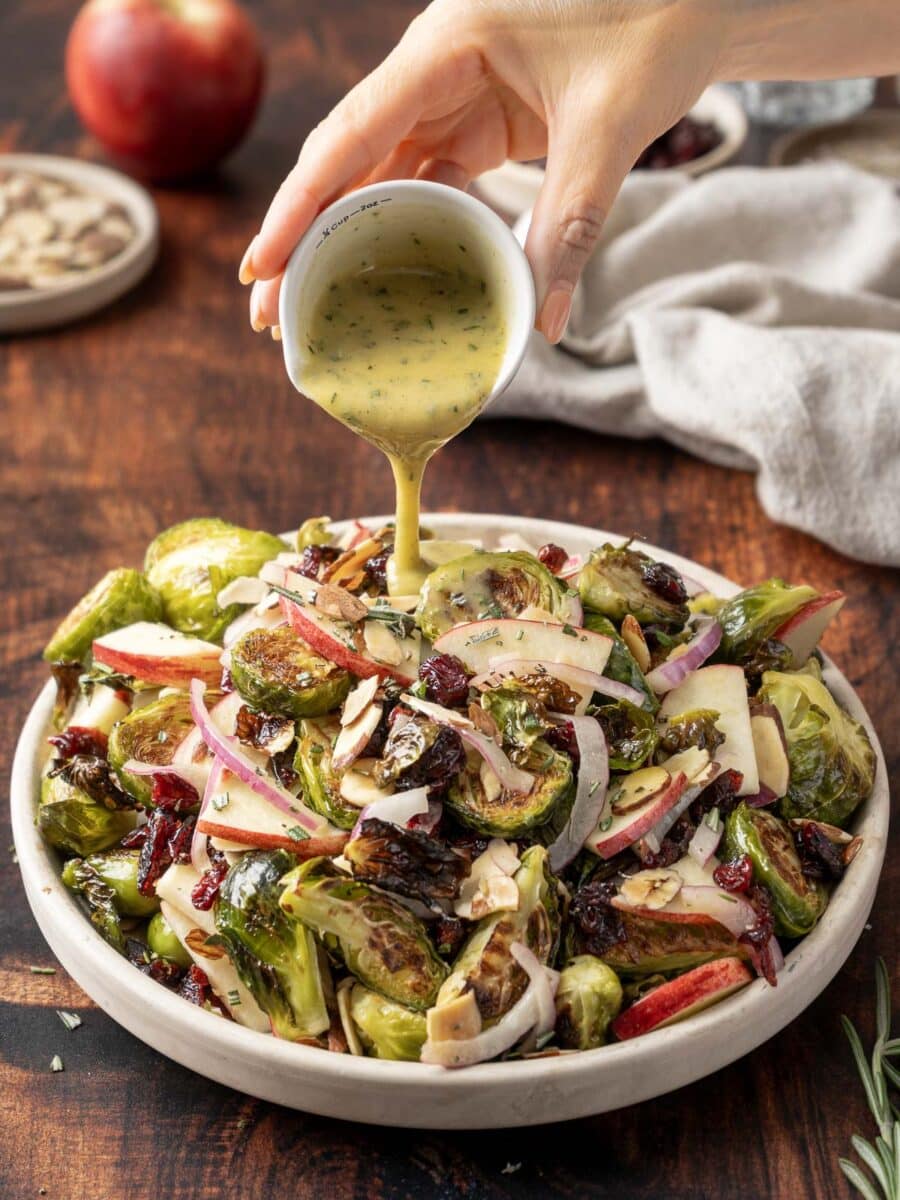 A hand pours dressing over a vibrant roasted brussels sprout salad with apples, cranberries, and sliced almonds on a wooden table. An apple and a cloth napkin linger in the background.