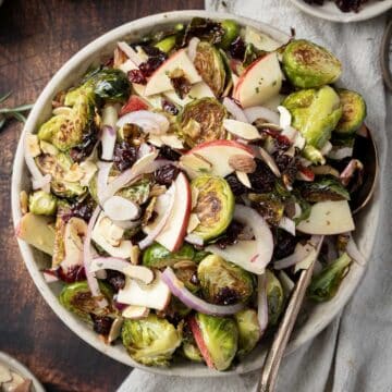 A bowl of roasted Brussels sprouts salad features sliced apples, almonds, cranberries, and red onions, served with a spoon.