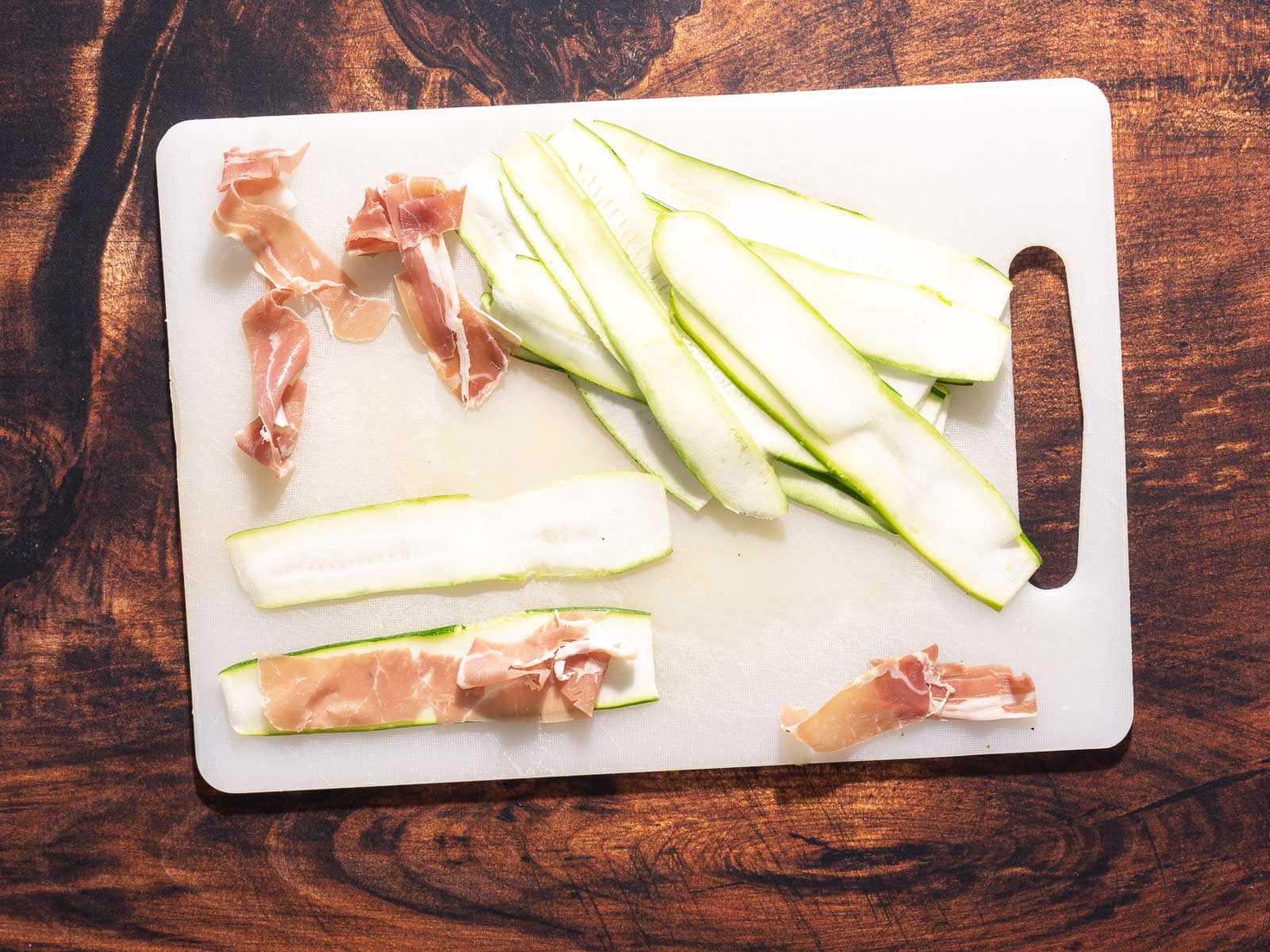 Zucchini appetizer featuring sliced zucchini and prosciutto laid neatly on a white cutting board, set atop a rustic wooden surface.