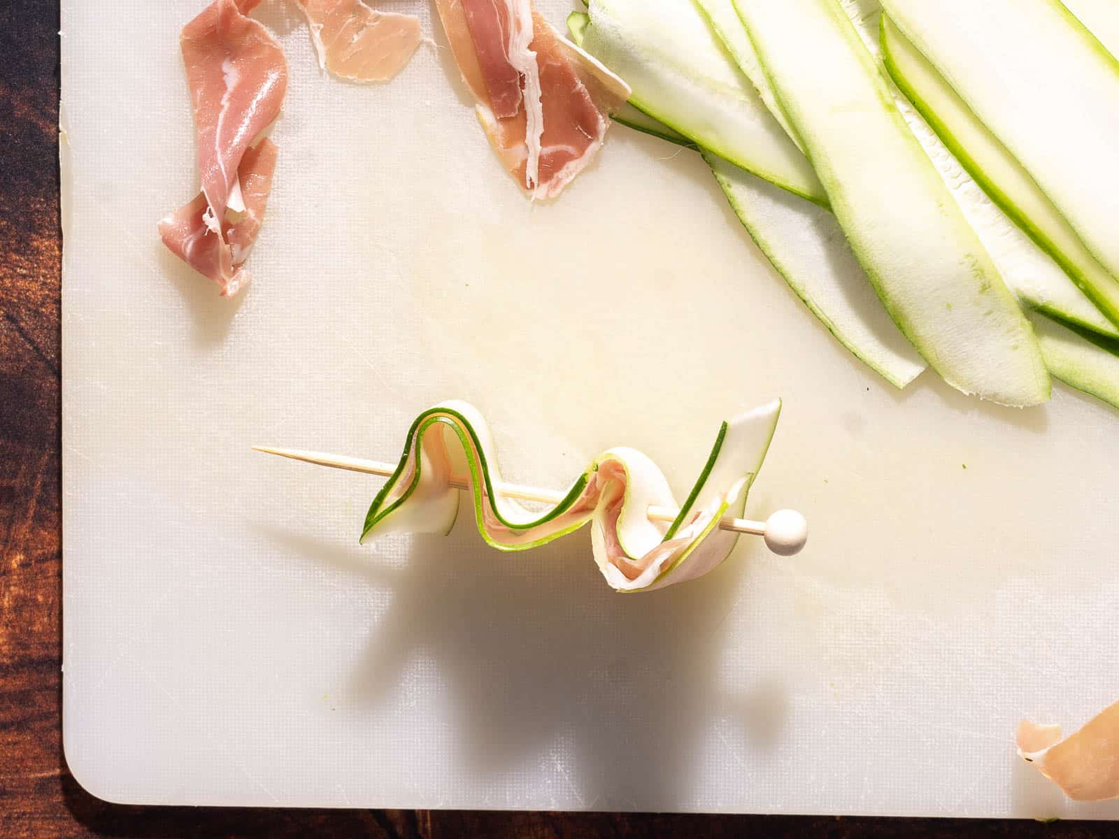 Zucchini appetizer skewers feature zucchini and prosciutto slices artfully threaded on a skewer, resting elegantly on a white cutting board with extra slices nearby.