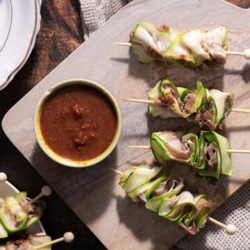 Enjoy our zucchini appetizer featuring skewers of rolled zucchini with tender meat, served alongside a bowl of rich red sauce on a rustic stone platter.