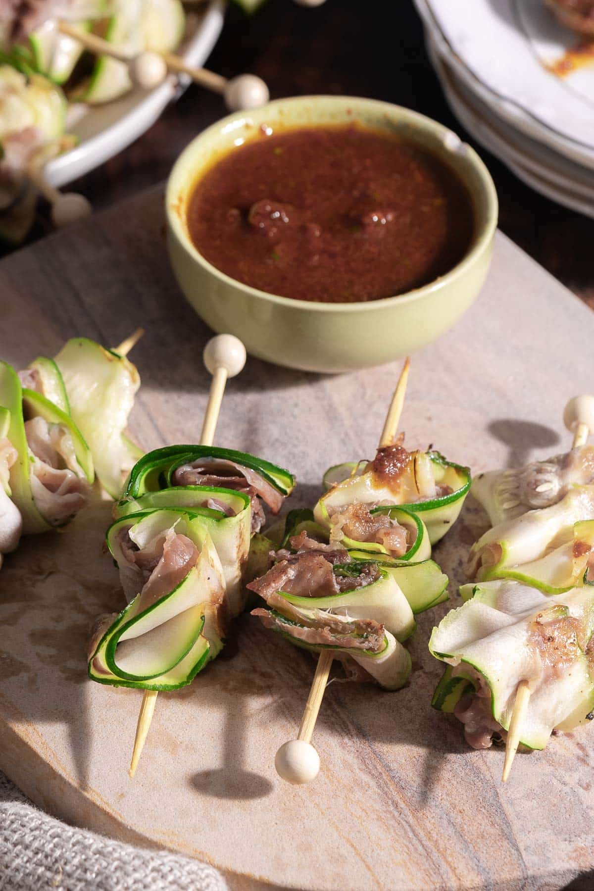 Delight in these skewered zucchini appetizer rolls with savory meat, perfectly paired next to a bowl of rich brown dipping sauce on a rustic wooden board.
