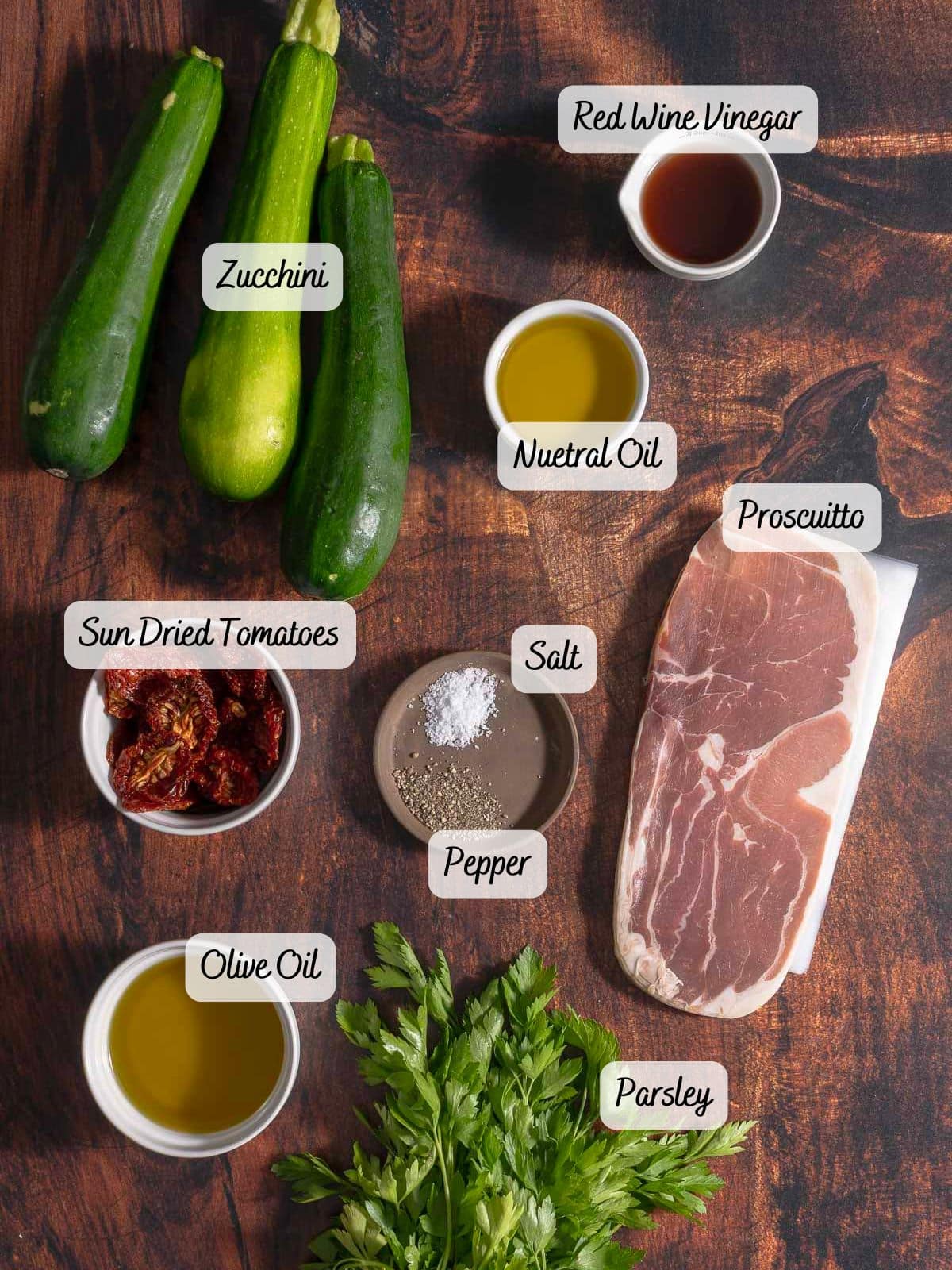 Ingredients laid out on a wooden surface: zucchini, red wine vinegar, prosciutto, sun-dried tomatoes, neutral oil, salt, pepper, olive oil, and parsley.