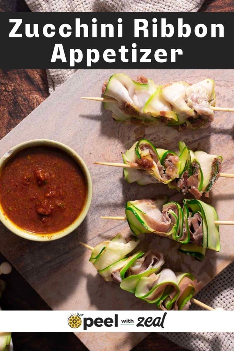 Zucchini ribbons and meat skewers on a wooden board with a bowl of red dipping sauce.
