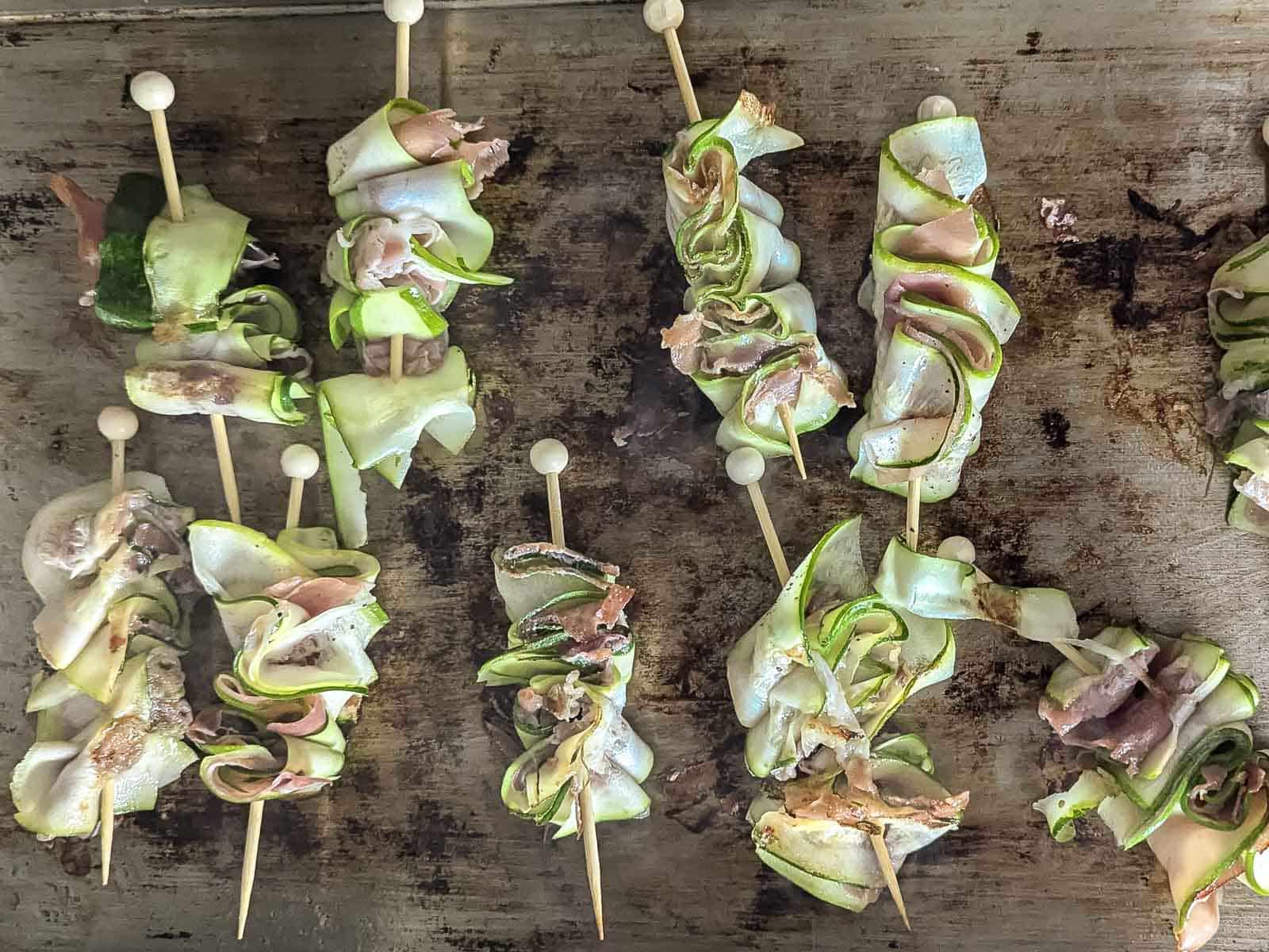 Skewered zucchini ribbons and prosciutto being seared on a flat top. 