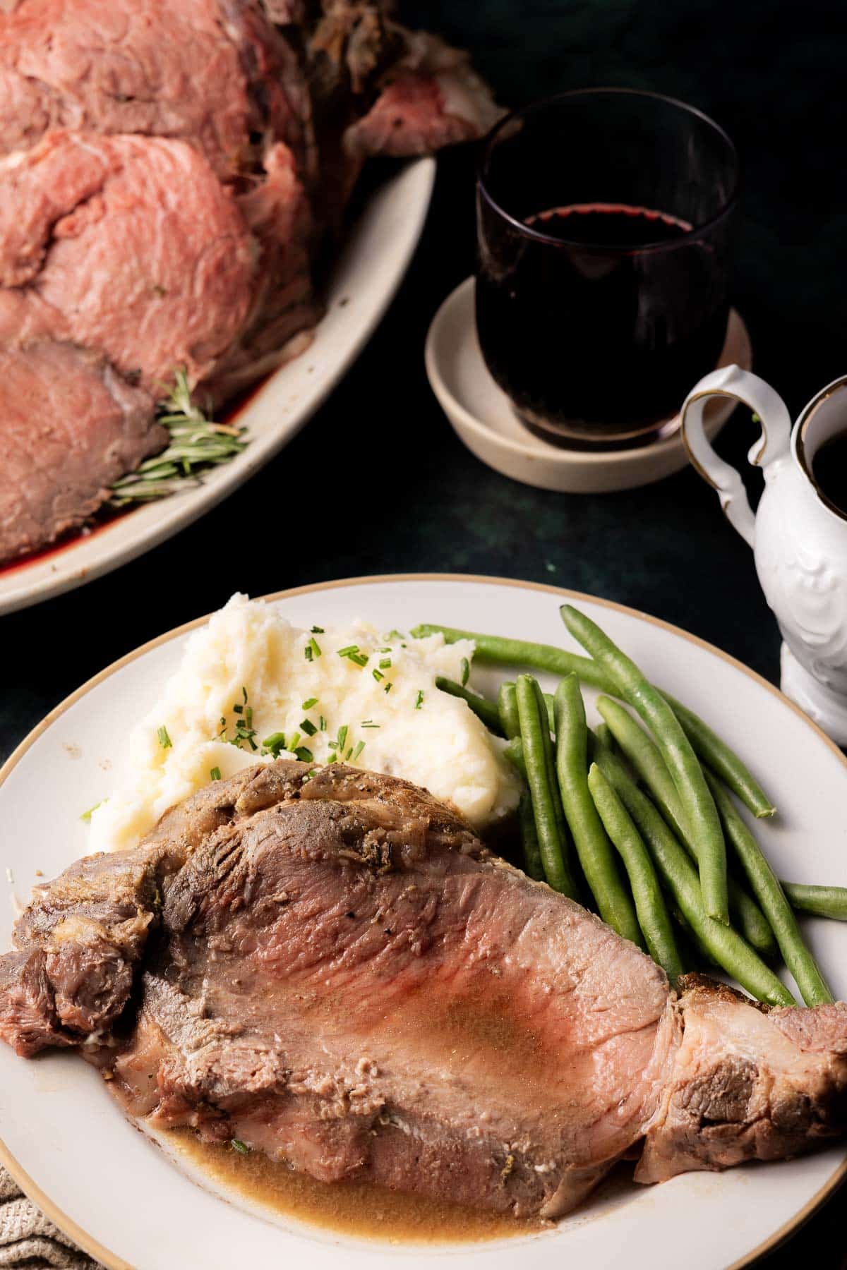 A plate of sliced roast beef, mashed potatoes with herbs, and green beans is complemented by a savory au jus. A glass of red wine and a gravy boat enhance the meal's richness.