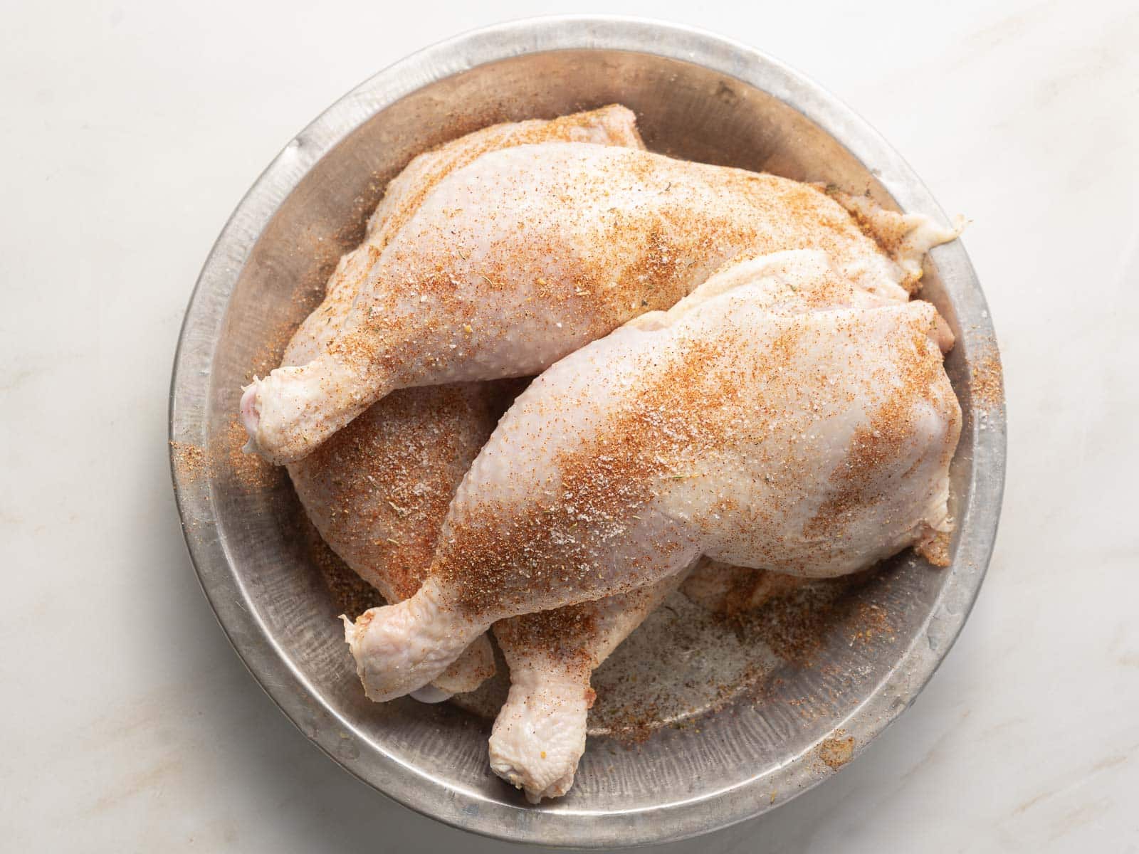 A metal bowl containing raw chicken legs, perfectly seasoned with Cajun spices, rests on a pristine white surface.