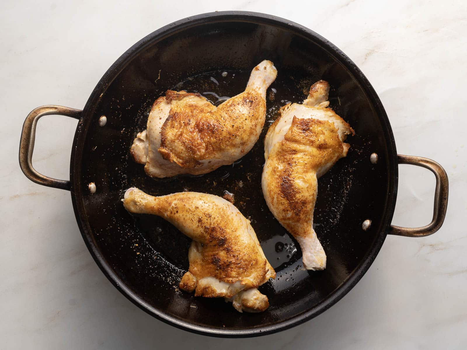 Three browned Cajun chicken legs sizzle in a black pan with metal handles, filling the air with a tantalizing aroma.