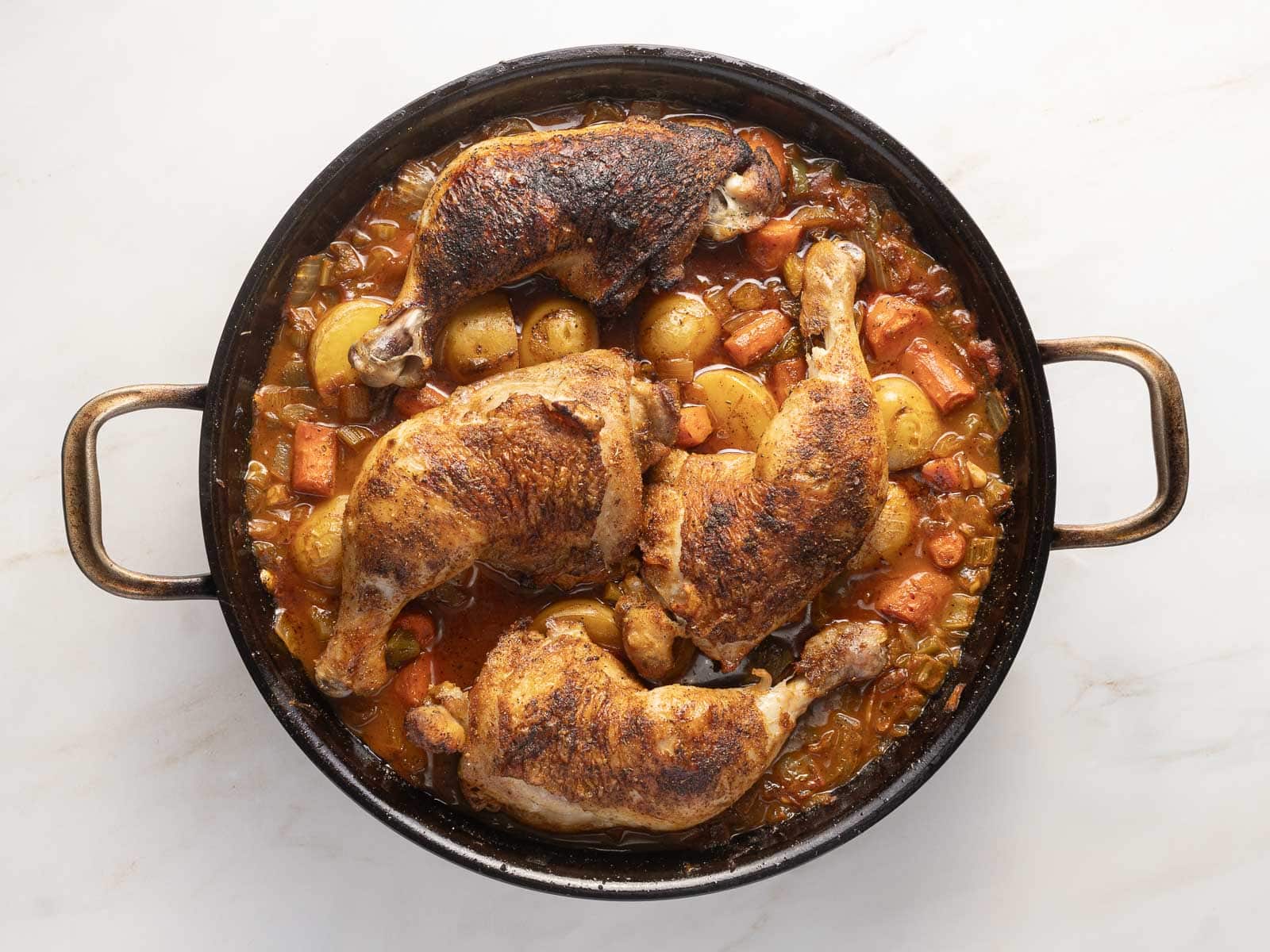 Roasted chicken legs with browned skin are nestled in a round pan, surrounded by potatoes, carrots, and onions in a delightful Cajun-inspired sauce.