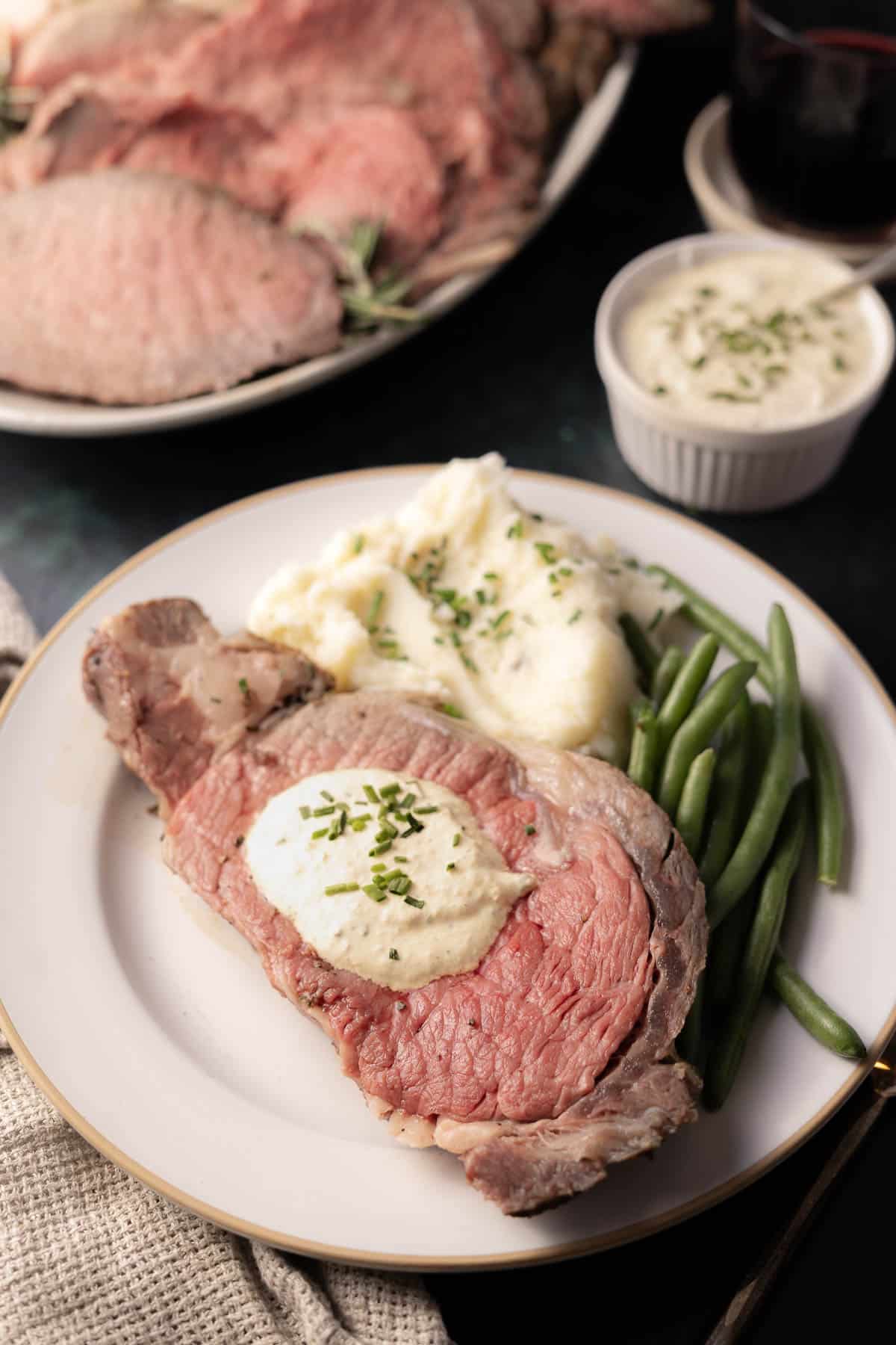 A plate with sliced roast beef topped with a creamy horseradish mustard sauce, accompanied by mashed potatoes and green beans. A sauce dish and an additional plate piled with more roast beef are subtly visible in the background.