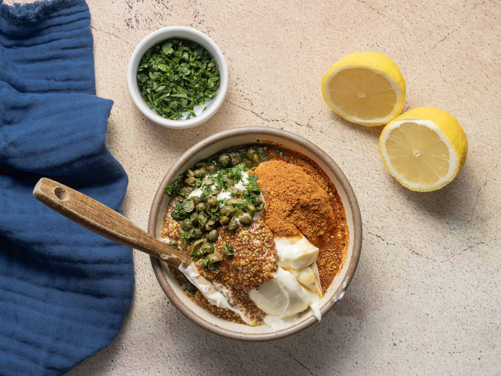A bowl with spices, capers, and a yogurt mix sits next to a wooden spoon, creating the perfect start for a delicious recipe. Nearby, a small bowl of chopped herbs and two lemon halves rest on a blue cloth on the textured surface.