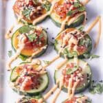 Cucumber slices topped with tuna tartare, drizzled with sauce, garnished with cilantro and sesame seeds, arranged on a white platter.