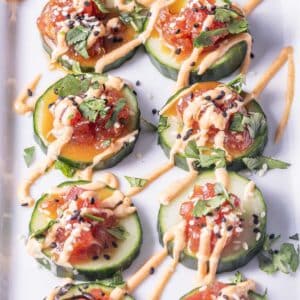 Cucumber slices topped with tuna tartare, drizzled with sauce, garnished with cilantro and sesame seeds, arranged on a white platter.