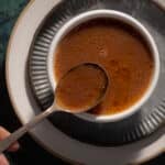 A cup of brown soup, rich and savory like au jus, sits on a plate with a spoon gracefully lifting the flavorful sauce.