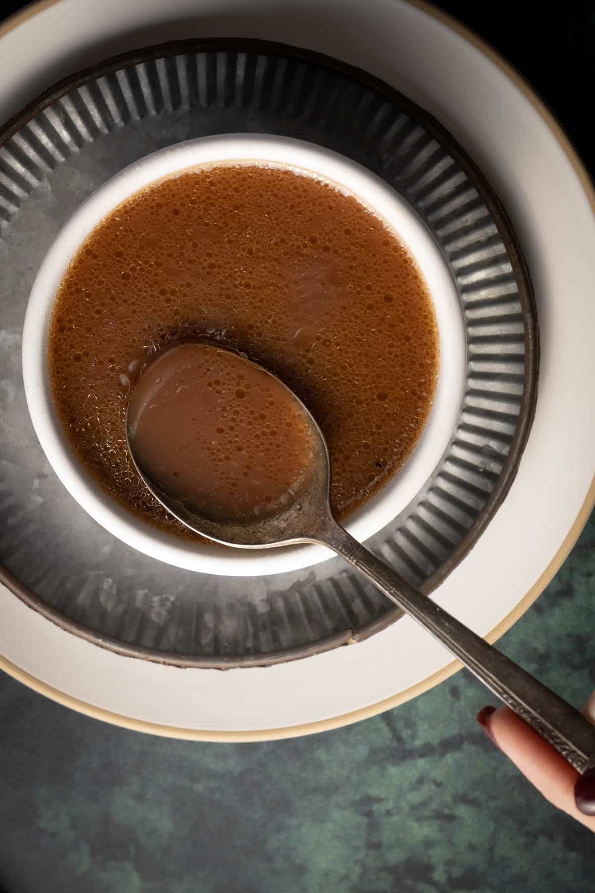 A spoon rests in a bowl of frothy brown soup, reminiscent of savory au jus, placed on a white and black plate. A hand with painted nails holds the bowl.