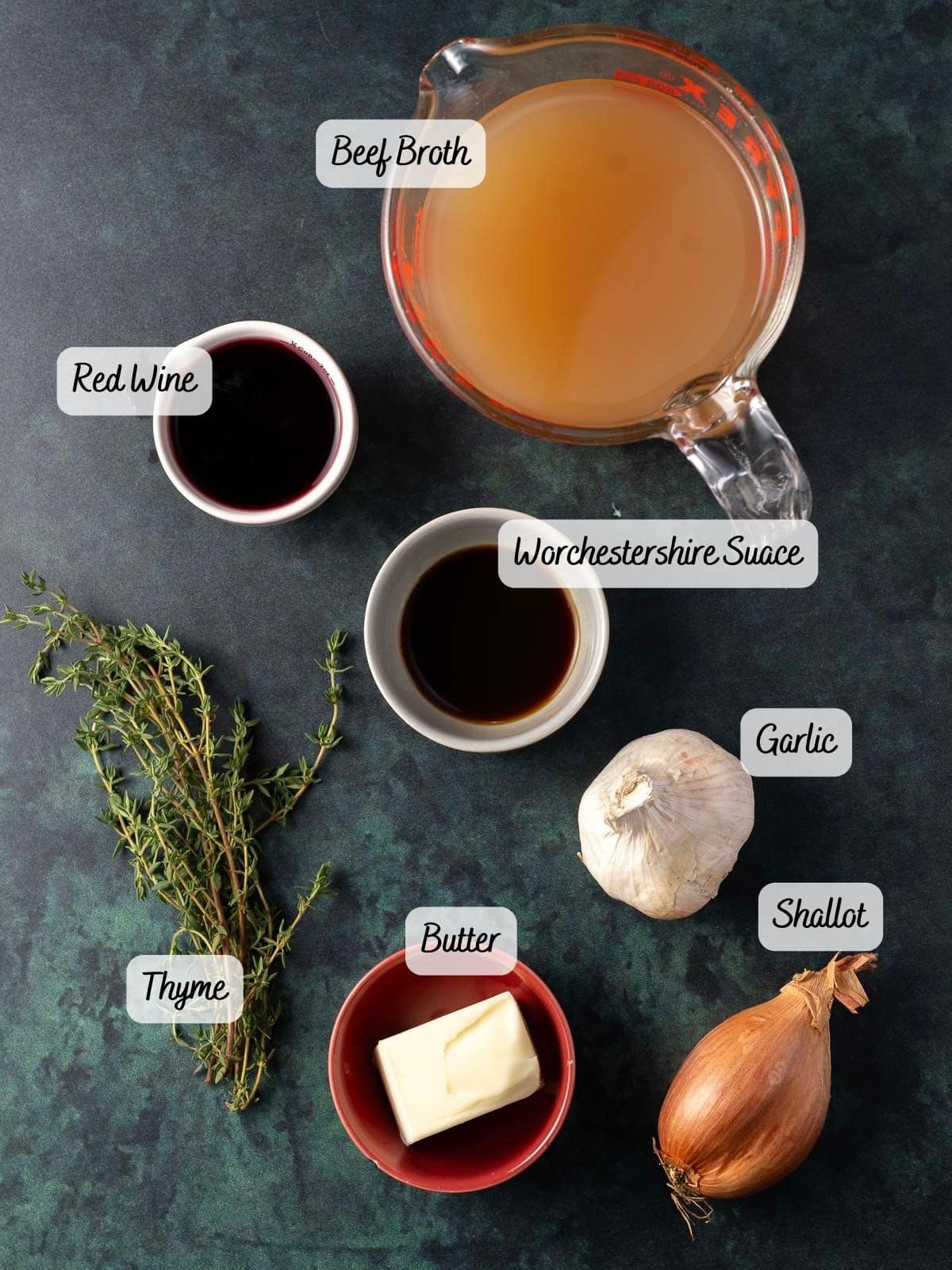 Ingredients for a rich au jus sauce on a dark surface: red wine, beef broth, Worcestershire sauce, garlic, shallot, butter, and thyme.