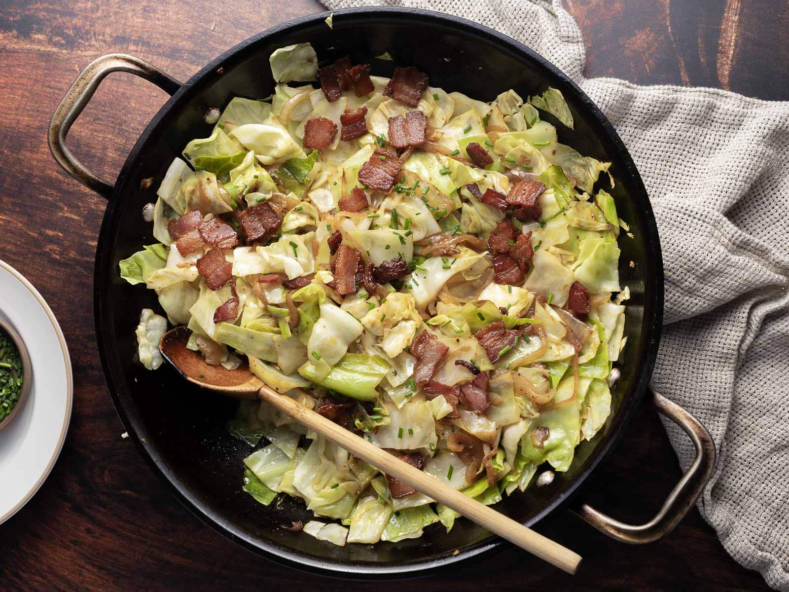 A skillet filled with fried cabbage and crispy bacon pieces rests on a wooden surface, accompanied by a wooden spoon. A beige cloth napkin is partially visible on the side, adding a rustic touch to this savory dish.