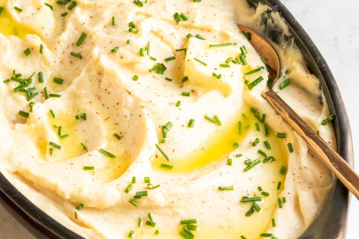 Mashed potatoes, the MVP of Thanksgiving sides, topped with chopped chives and a drizzle of melted butter, served in a black dish with a spoon.