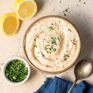 A bowl of creamy dip, perfect for any feast, is garnished with chopped herbs alongside lemon halves and a small bowl of more herbs. A spoon rests nearby on a blue napkin, complementing this delightful recipe.