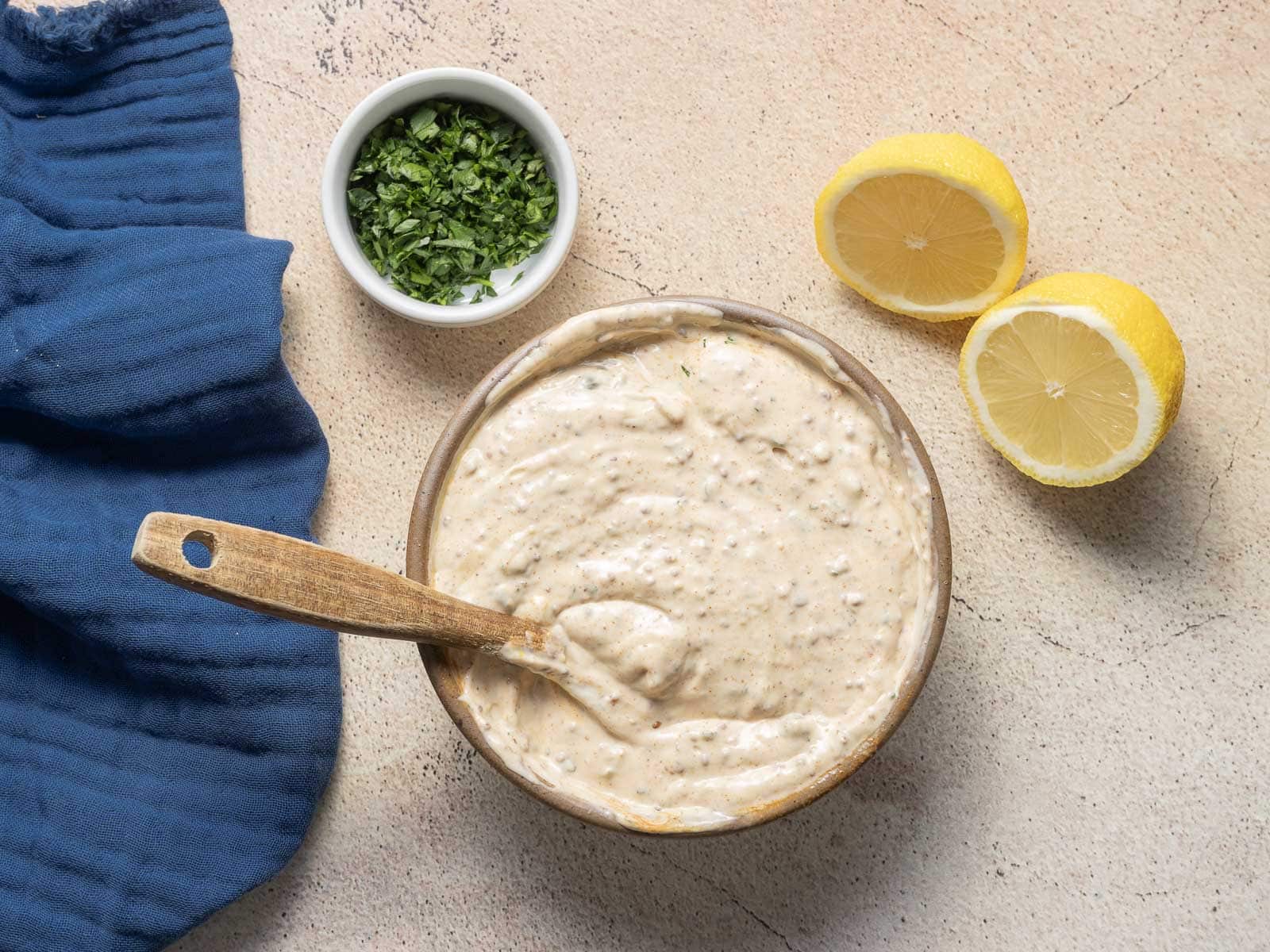 A bowl of creamy dip with a wooden spoon rests on a textured surface, surrounded by a blue cloth and a small dish of chopped herbs. Two lemon halves add zest to this feast, perfect for any gathering or recipe inspiration.