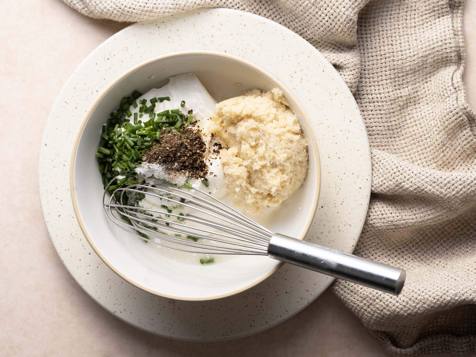 A bowl contains chopped chives, black pepper, salt, mayonnaise, and grated cheese with a hint of horseradish sauce. A whisk rests on the edge of the bowl. A textured cloth is in the background.