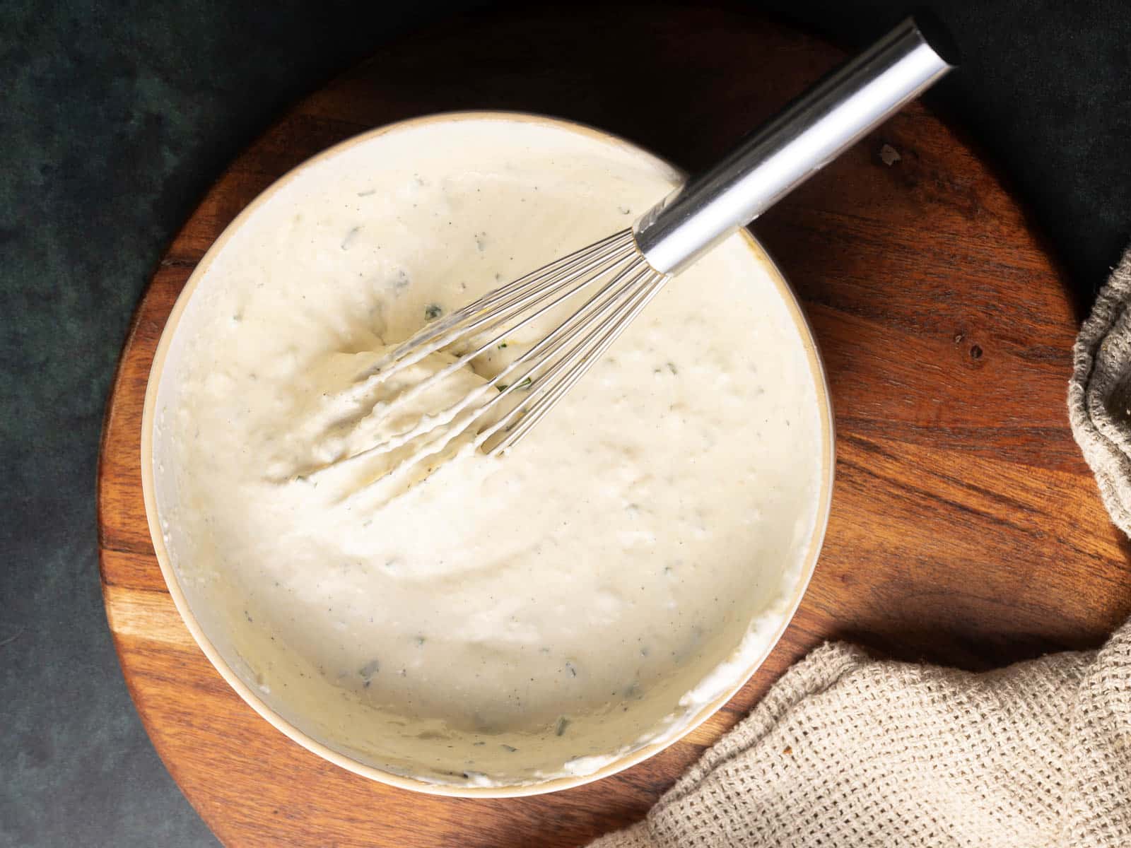 A bowl of creamy horseradish mustard sauce with herbs mixed in, shown with a metal whisk on a wooden board, next to a textured cloth.