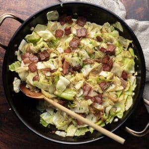 A skillet of fried cabbage with crispy bacon pieces and tender onions, accompanied by a wooden spoon on a wooden surface.