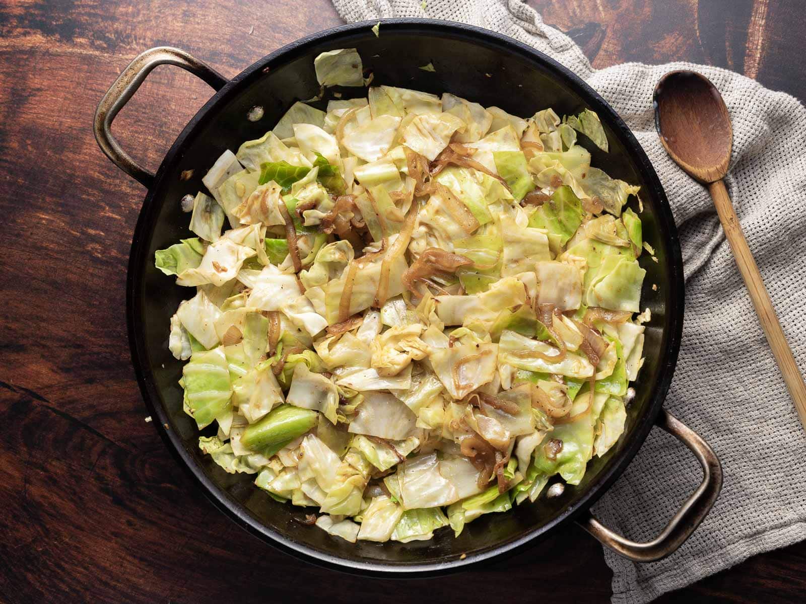 A skillet filled with fried cabbage, crispy bacon, and saut&eacute;ed onions rests on a wooden surface, accompanied by a wooden spoon and a gray cloth.