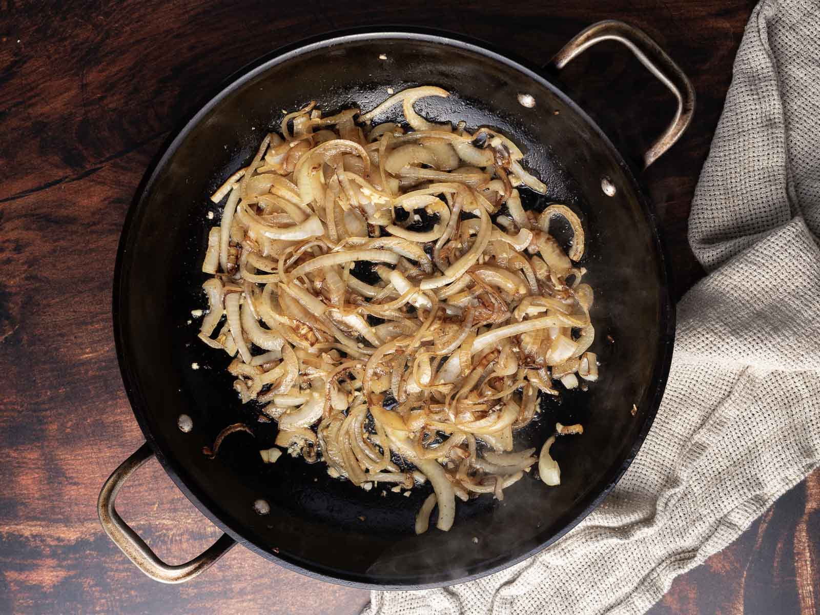 Sliced onions caramelizing in a black pan on a wooden surface, with a cloth nearby.