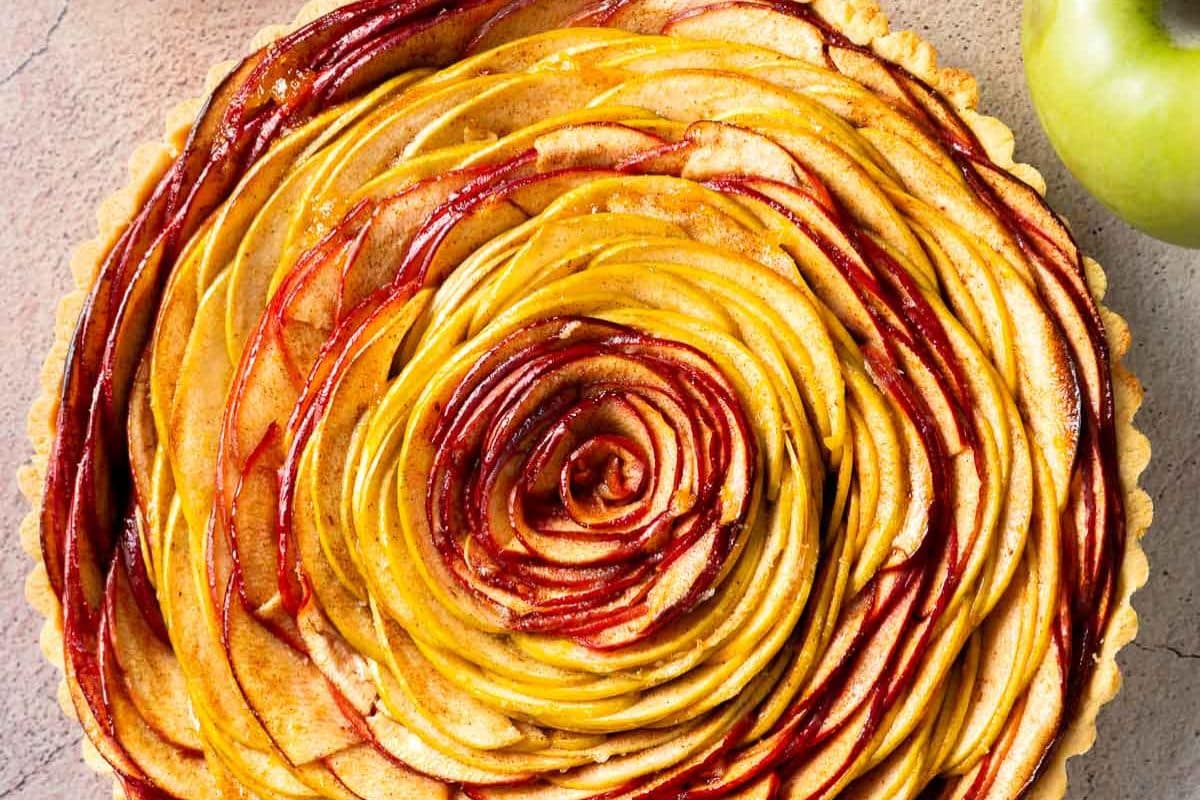 A close-up of a spiral apple tart with thinly sliced red and yellow apples arranged in a circular pattern. Perfect for Thanksgiving, this gluten-free dessert showcases an artful presentation, with an apple partially visible beside it.